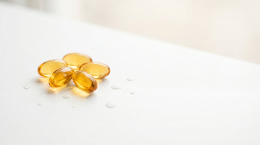 Cápsulas softgel douradas de ômega 3 com gotas de água sobre superfície branca — guia 3VS Nutrition